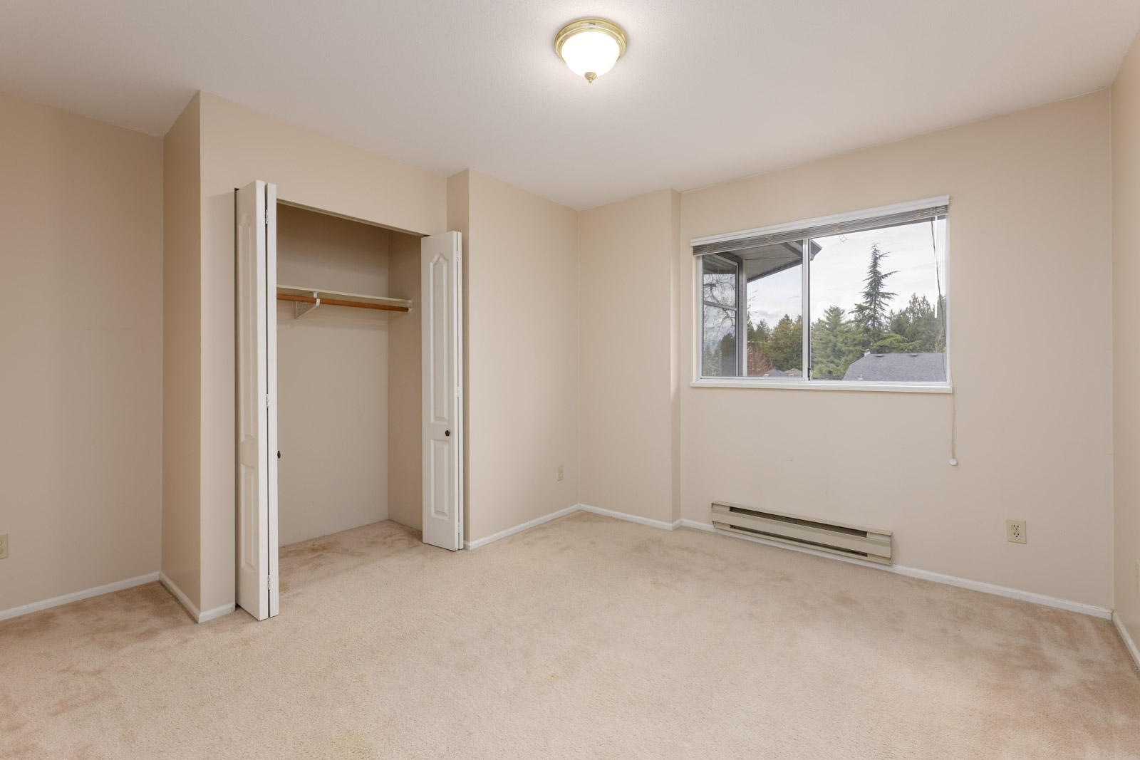 Empty beige room with a closed door, open closet, single window, and baseboard heater. The floor is carpeted, and a ceiling light fixture is present.