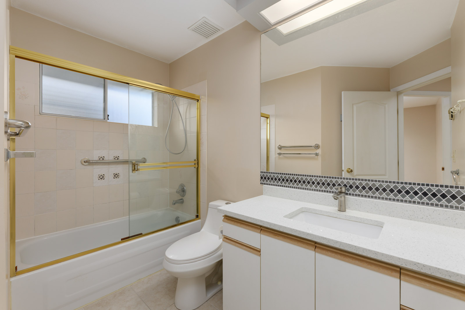 Bathroom with a glass-enclosed bathtub, beige tiles, and a large mirror above a sink with white countertop. Toilet beside the vanity, and a door partially visible.