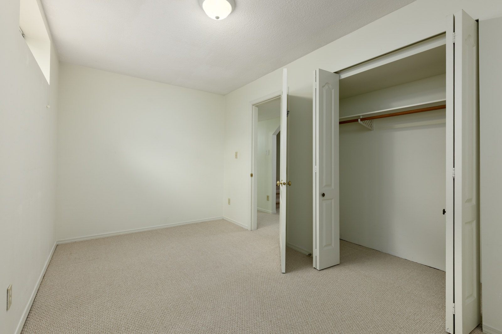 Empty room with beige carpet, white walls, and a ceiling light. An open door reveals a closet with a wooden rod and shelf. Another door leads out of the room.