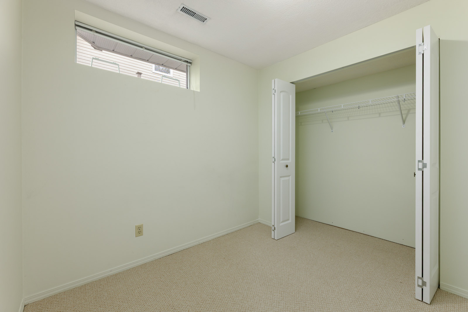 A small, empty room with light green walls, a vent, a small window near the ceiling, an open closet with a white folding door, and beige carpet flooring.
