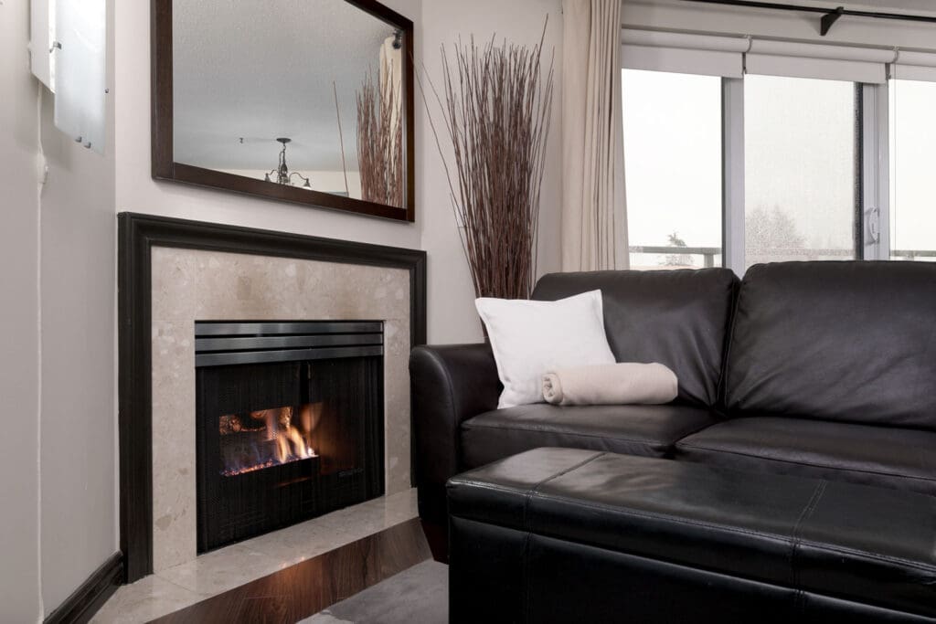 Modern living room with a lit fireplace, black leather sofa, white pillow, and blanket. Large window with curtains and decorative branches in a corner.