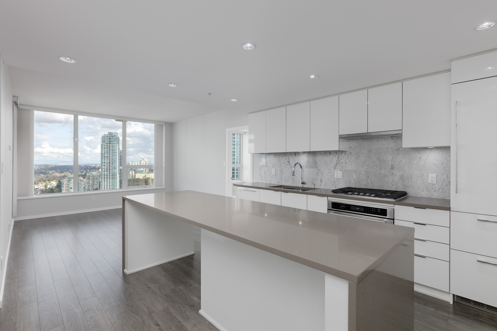 Modern apartment kitchen with white cabinets, gray countertop island, built-in appliances, and large windows offering a city view.