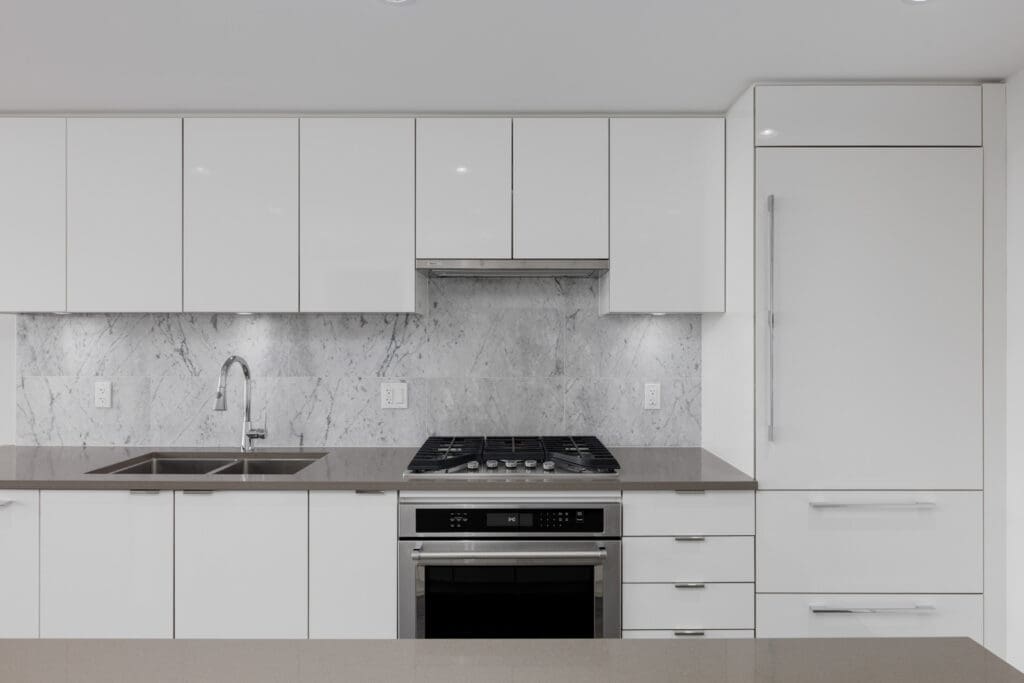 Modern kitchen with white cabinets, stainless steel oven and stove, double sink, marble backsplash, and integrated refrigerator.