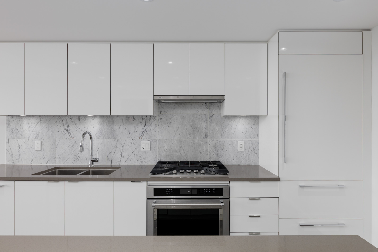 Modern kitchen with white cabinets, stainless steel oven and stove, double sink, marble backsplash, and integrated refrigerator.