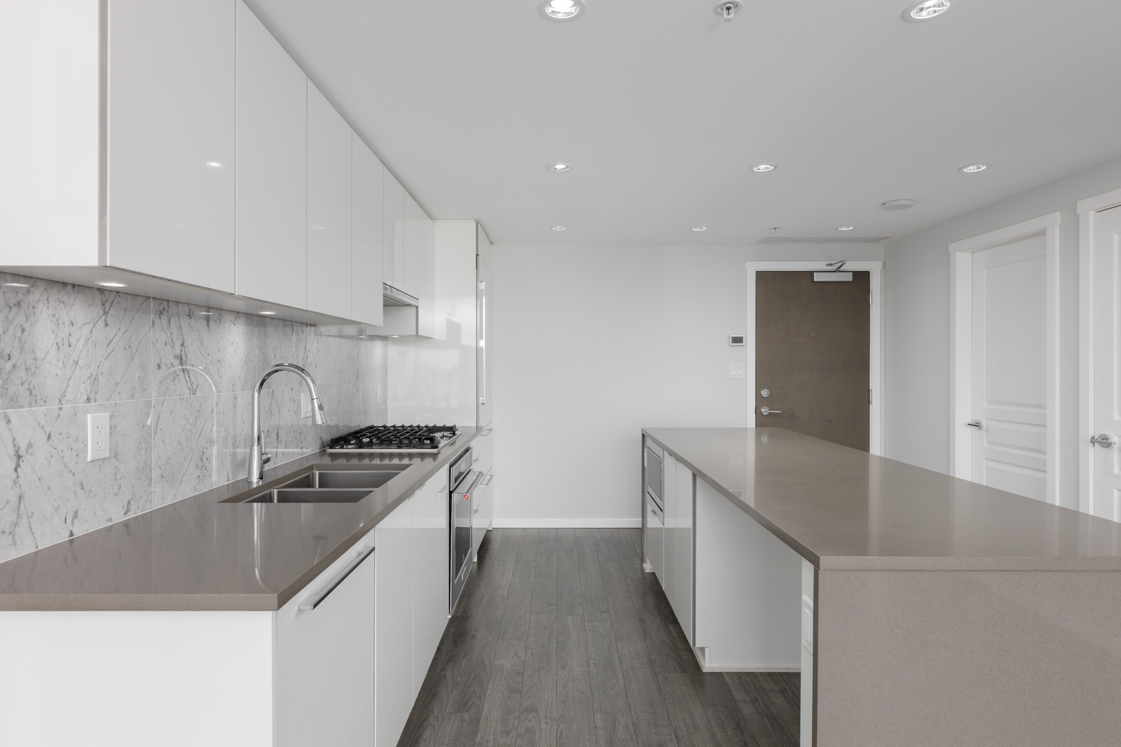 Modern kitchen with white cabinets, marble backsplash, stainless steel appliances, double sink, and a large island with a gray countertop on dark wood flooring.