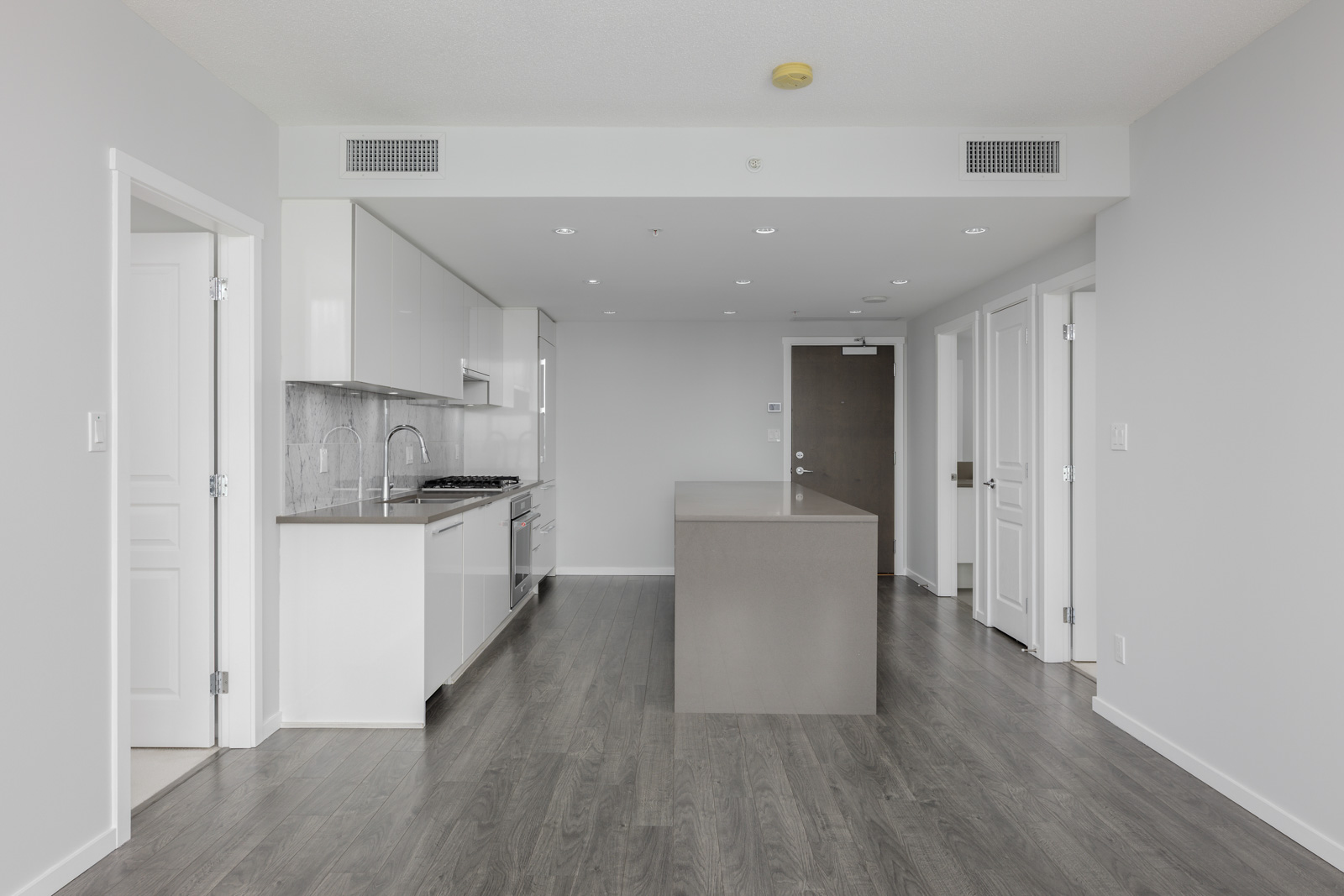 Modern kitchen with white cabinets, gray countertops, central island, stainless steel appliances, and wood-look flooring in a bright, empty apartment.