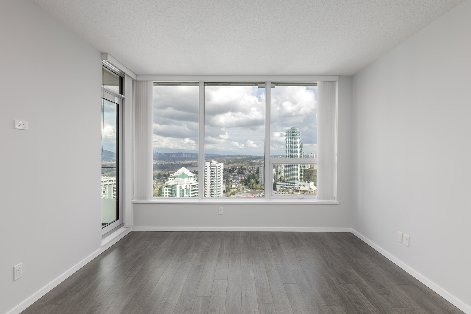 Empty room with gray wood flooring, white walls, and large windows offering a cityscape and mountain view under a cloudy sky.