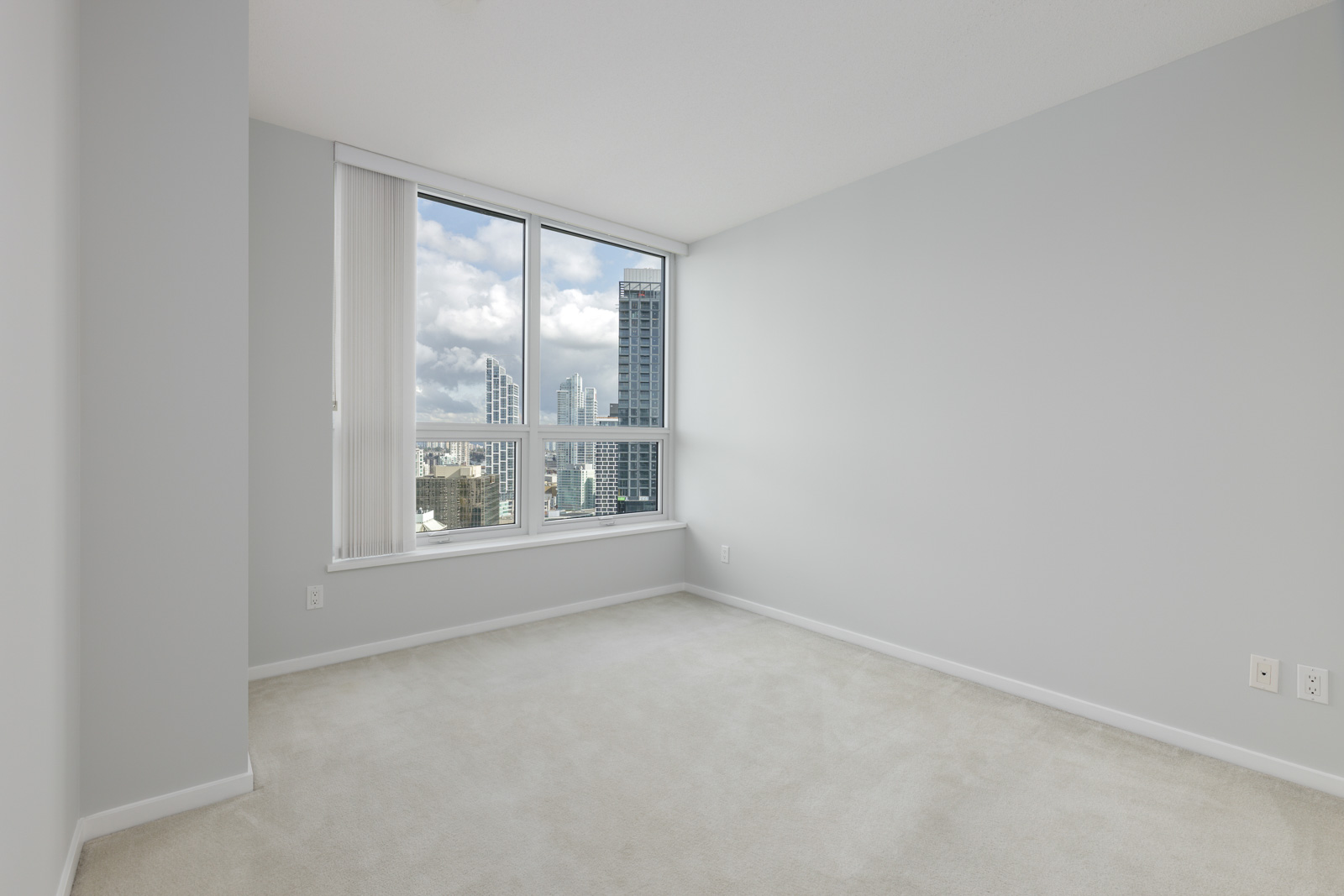 Empty room with light gray walls and carpet, large window showing city buildings and a cloudy sky outside.