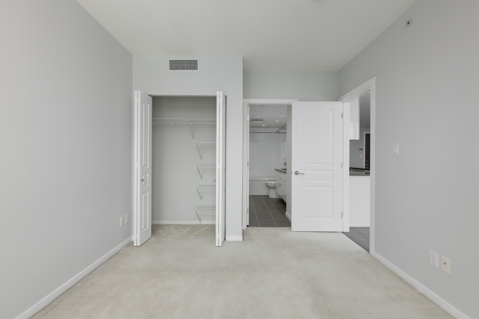 Empty bedroom with light gray walls, beige carpet, open closet with shelves, and open doors revealing a bathroom and hallway.