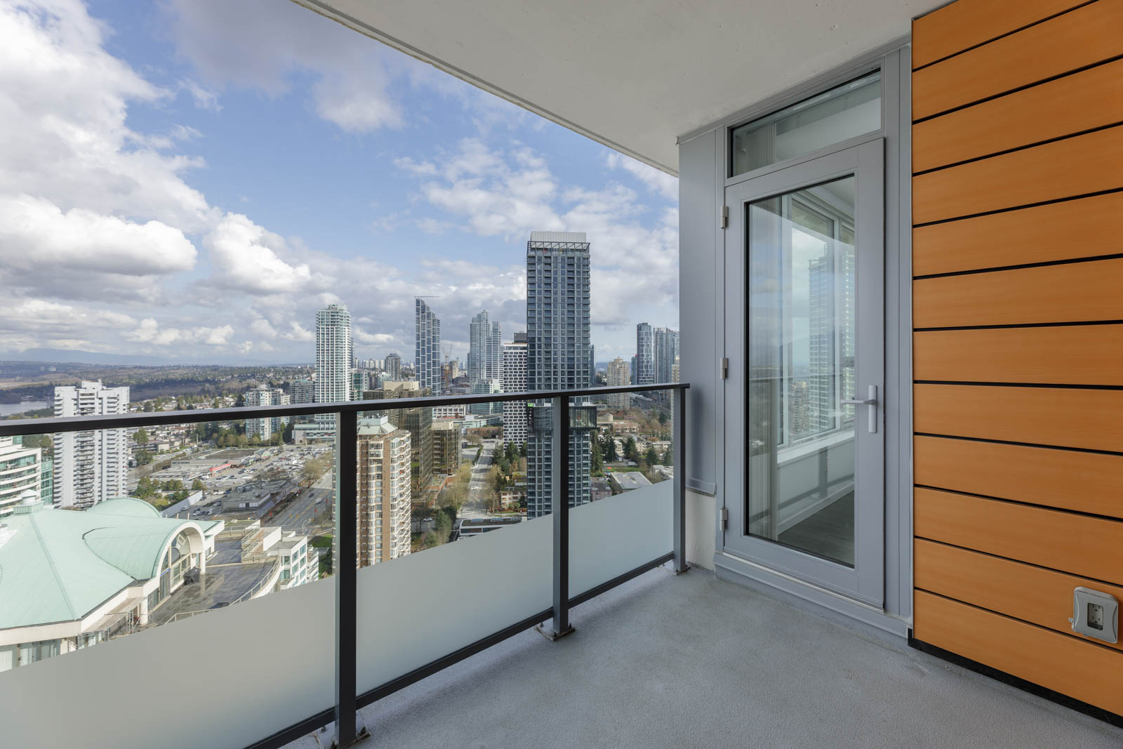 High-rise balcony with glass railing overlooking a cityscape of tall buildings under a partly cloudy sky.