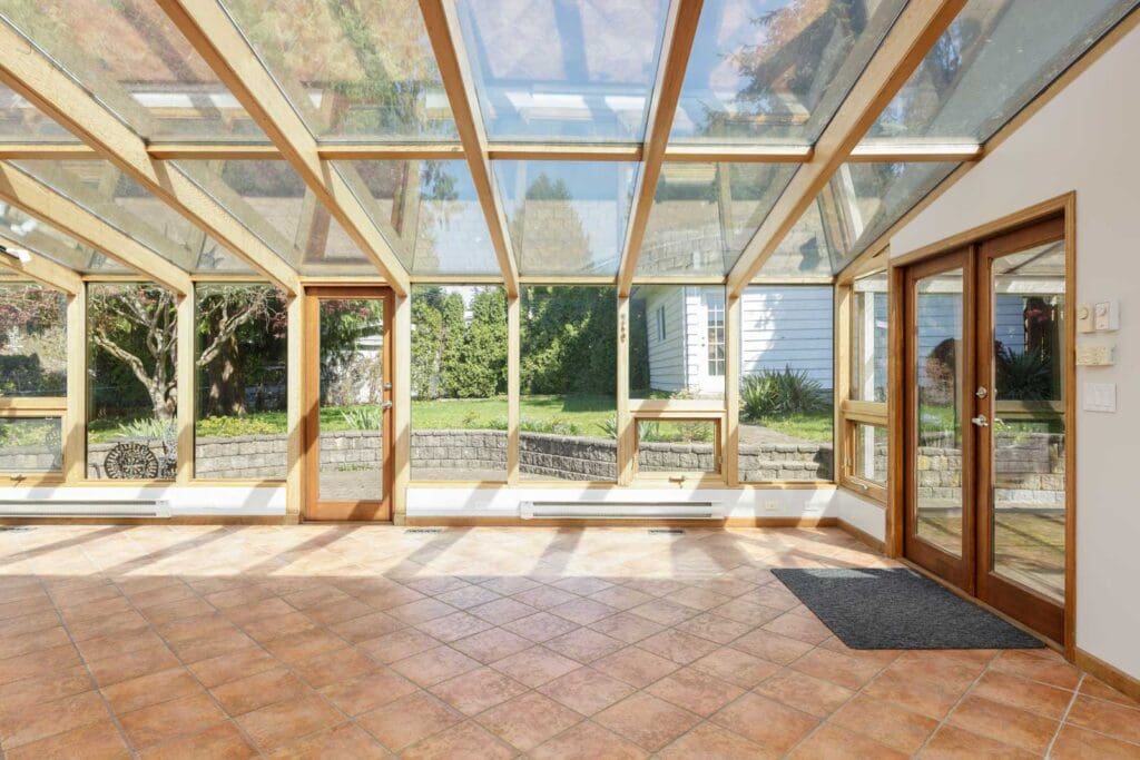 A sunroom with a glass ceiling and walls, tile flooring, and wooden-framed doors and windows, overlooking a backyard garden.