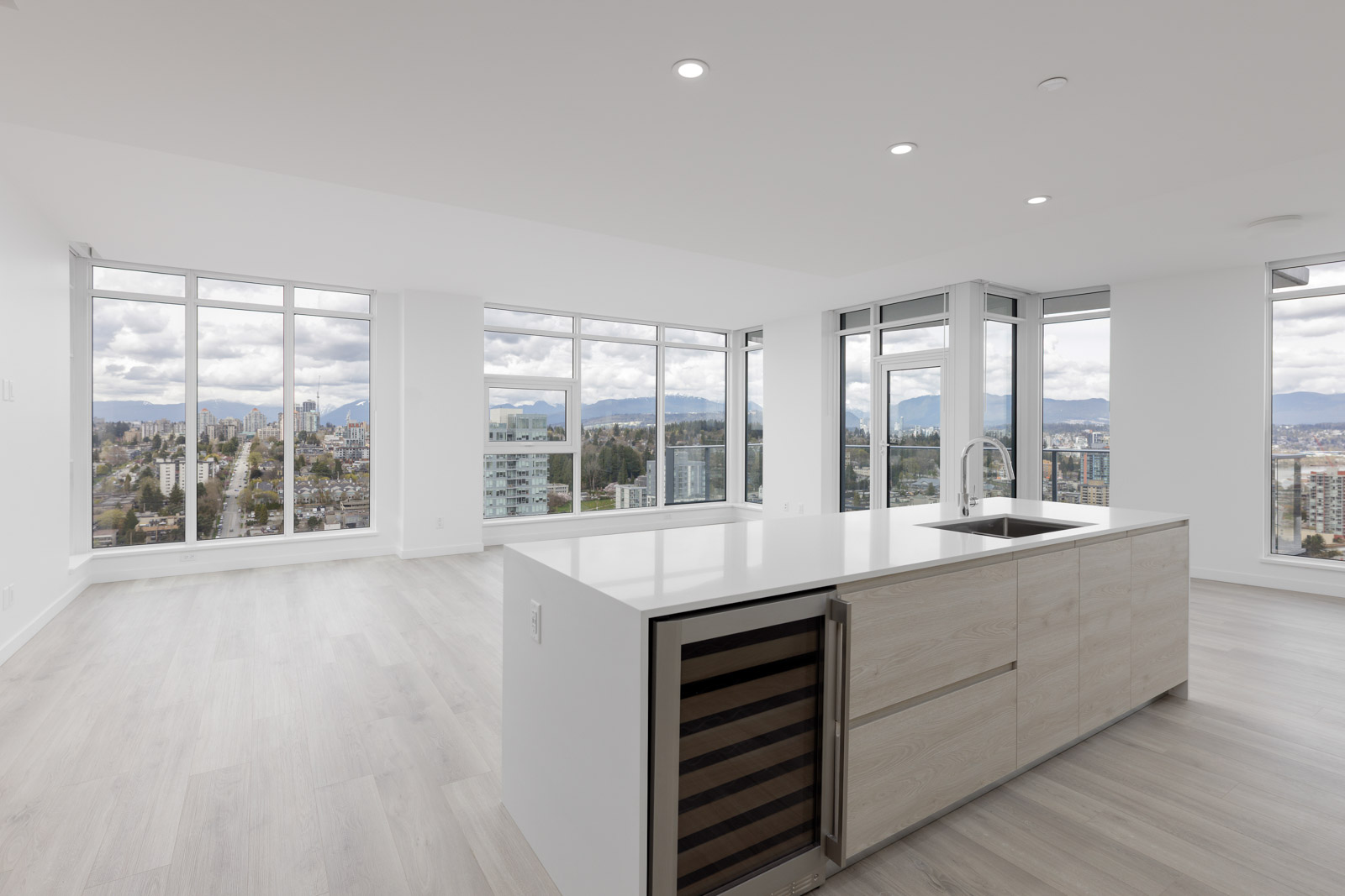 Modern, empty apartment with large windows, light wood floors, an island counter with a sink, and a built-in wine cooler. City skyline and mountains visible through the windows.