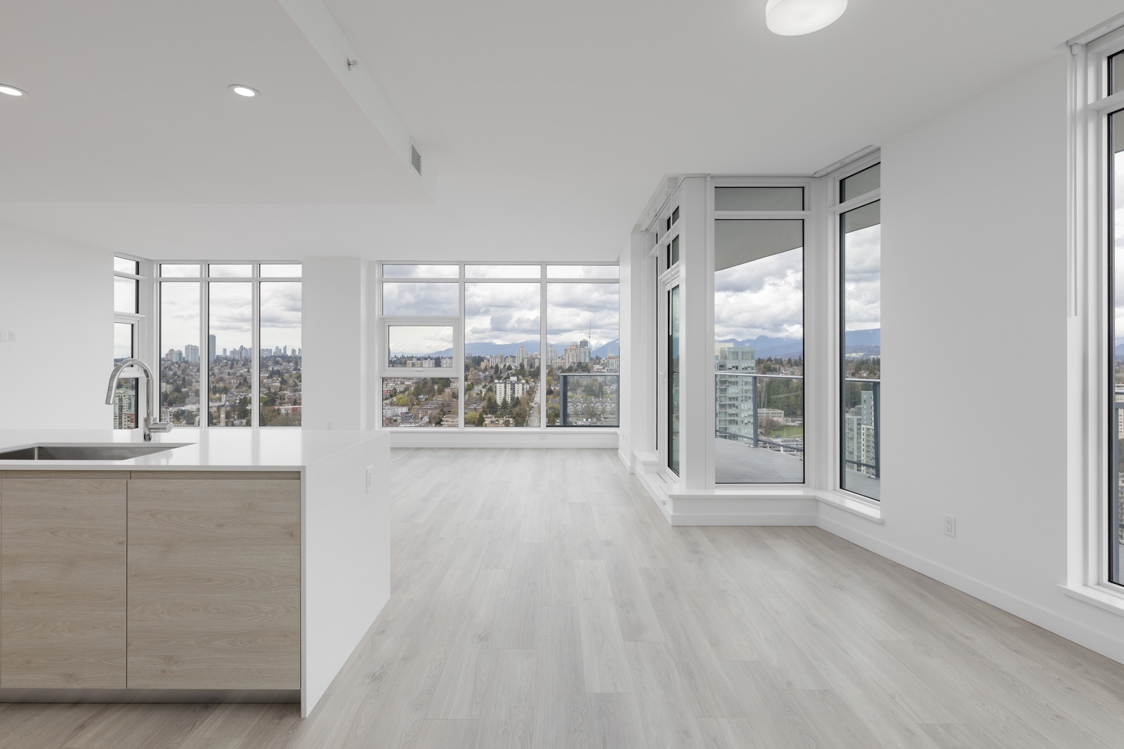 A modern, unfurnished apartment with large windows, offering a panoramic city skyline view. The room features light wood flooring and white walls, with an island kitchen in the foreground.