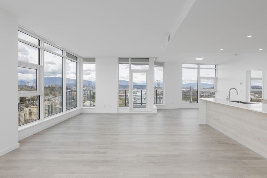 Spacious empty apartment with large windows, light wood flooring, and a modern kitchen, offering panoramic city and mountain views.