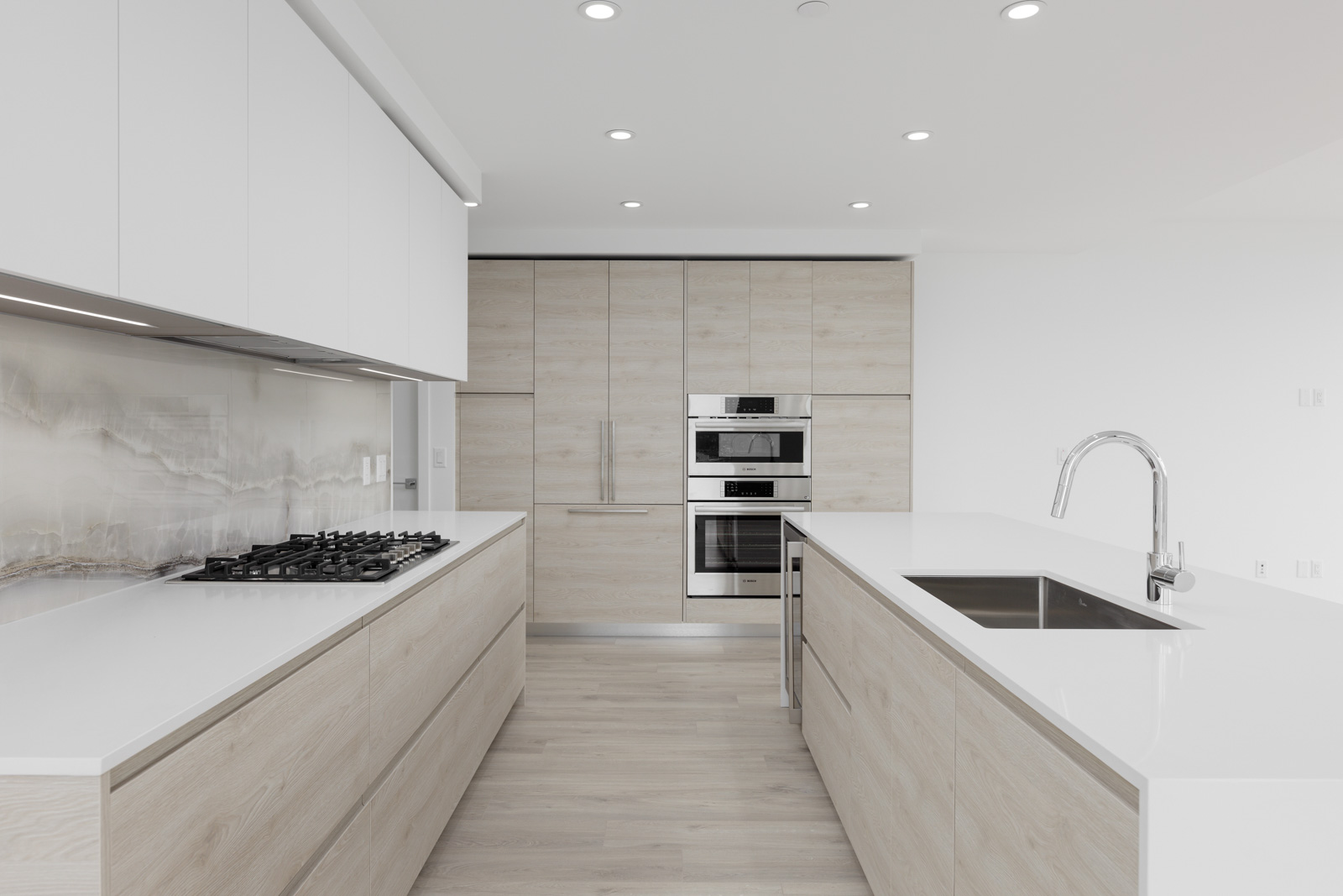 Minimalist kitchen with light wood cabinets, a gas stovetop, wall ovens, and a sleek island featuring a sink with a curved faucet. Bright and modern design with recessed lighting.