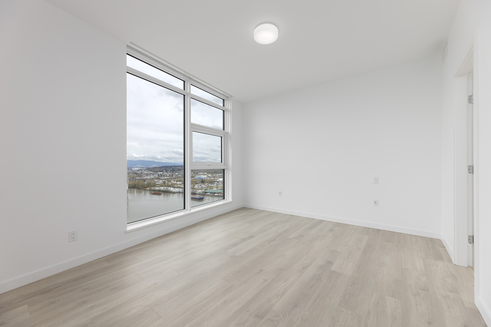 Empty room with light wood flooring, white walls, and a large window with a view of a river and mountains. Ceiling light fixture is turned on.