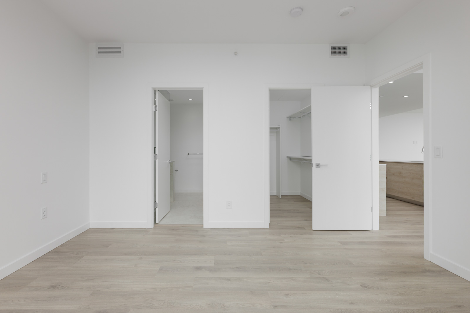 Empty room with white walls and light wood flooring. Two open doors lead to a closet and a bathroom. Ceiling vents and lights are present.