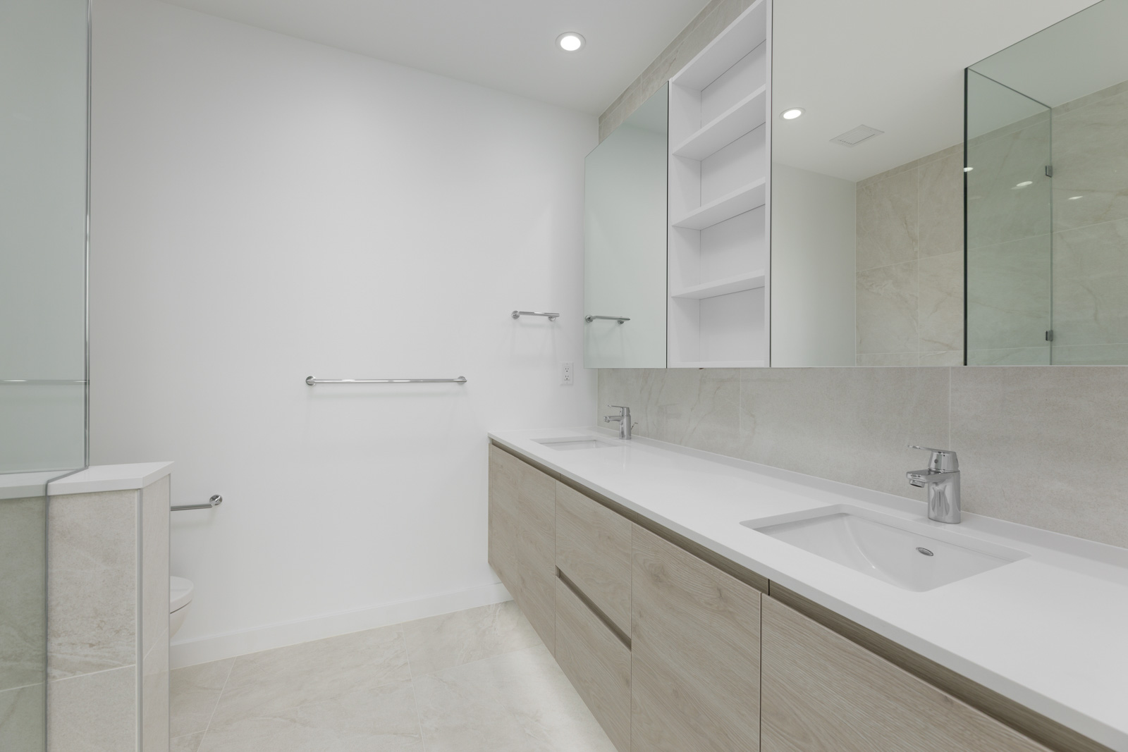 Modern bathroom with beige tile flooring and walls, featuring a large mirror, two sinks with chrome faucets, white cabinets, and a towel rack.
