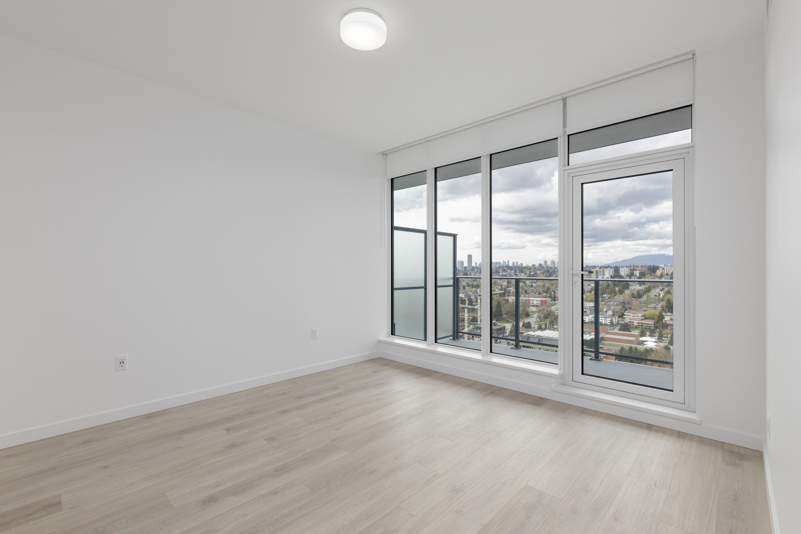 Empty room with light wood flooring, white walls, and large windows offering a view of a city skyline and overcast sky. A door to the balcony is partially open.
