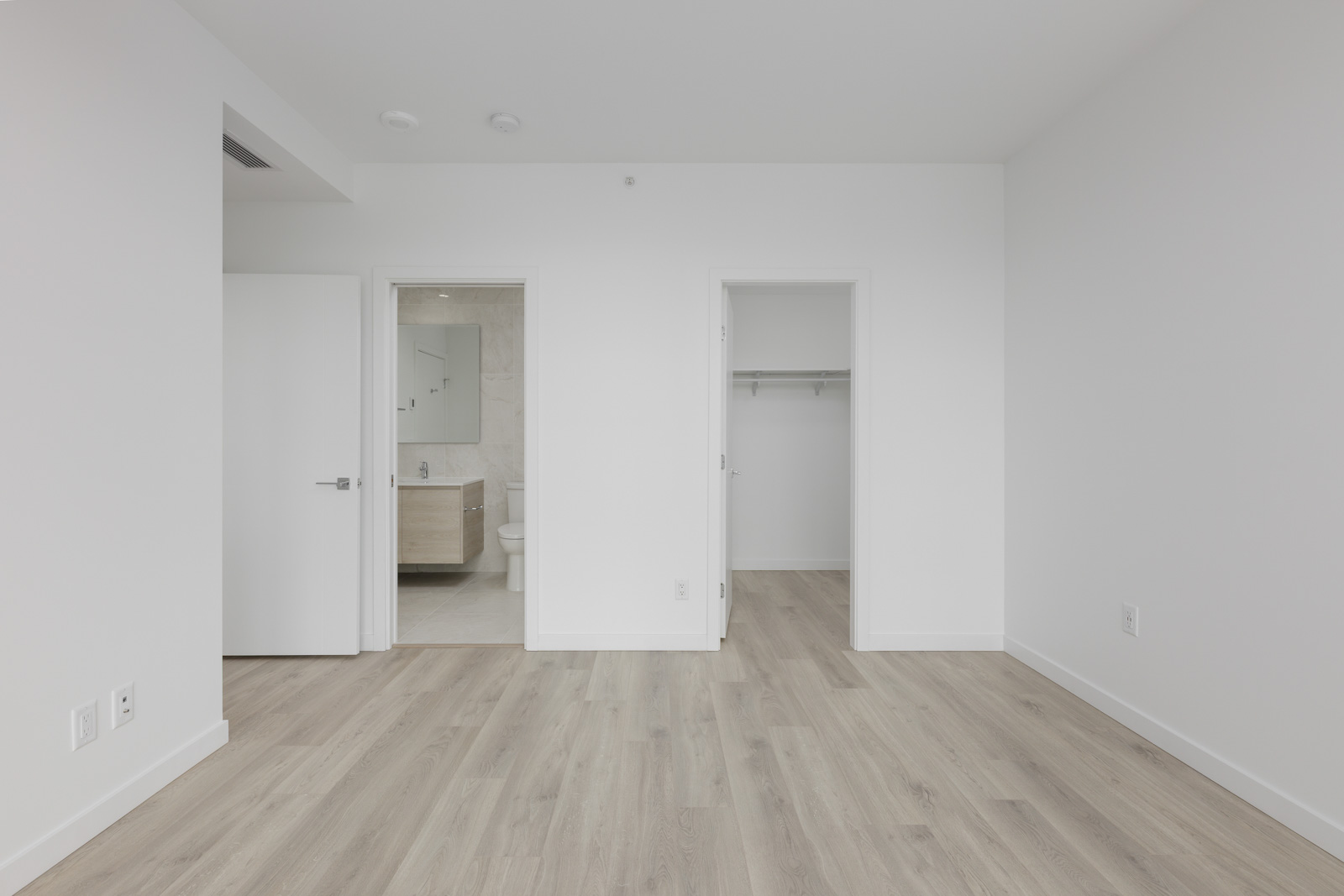 Empty room with light wood flooring, white walls, and two open doors. One door leads to a bathroom with a visible sink and mirror. The other door opens to a small closet.