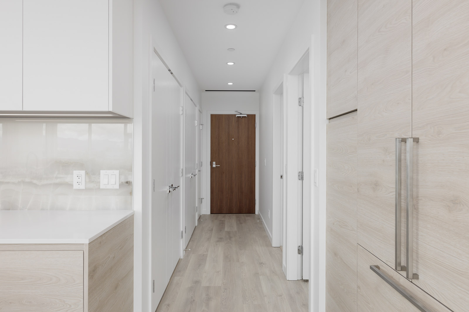 Minimalist hallway with light wood flooring, white walls, and a wooden front door. Modern cabinetry is visible on the right.