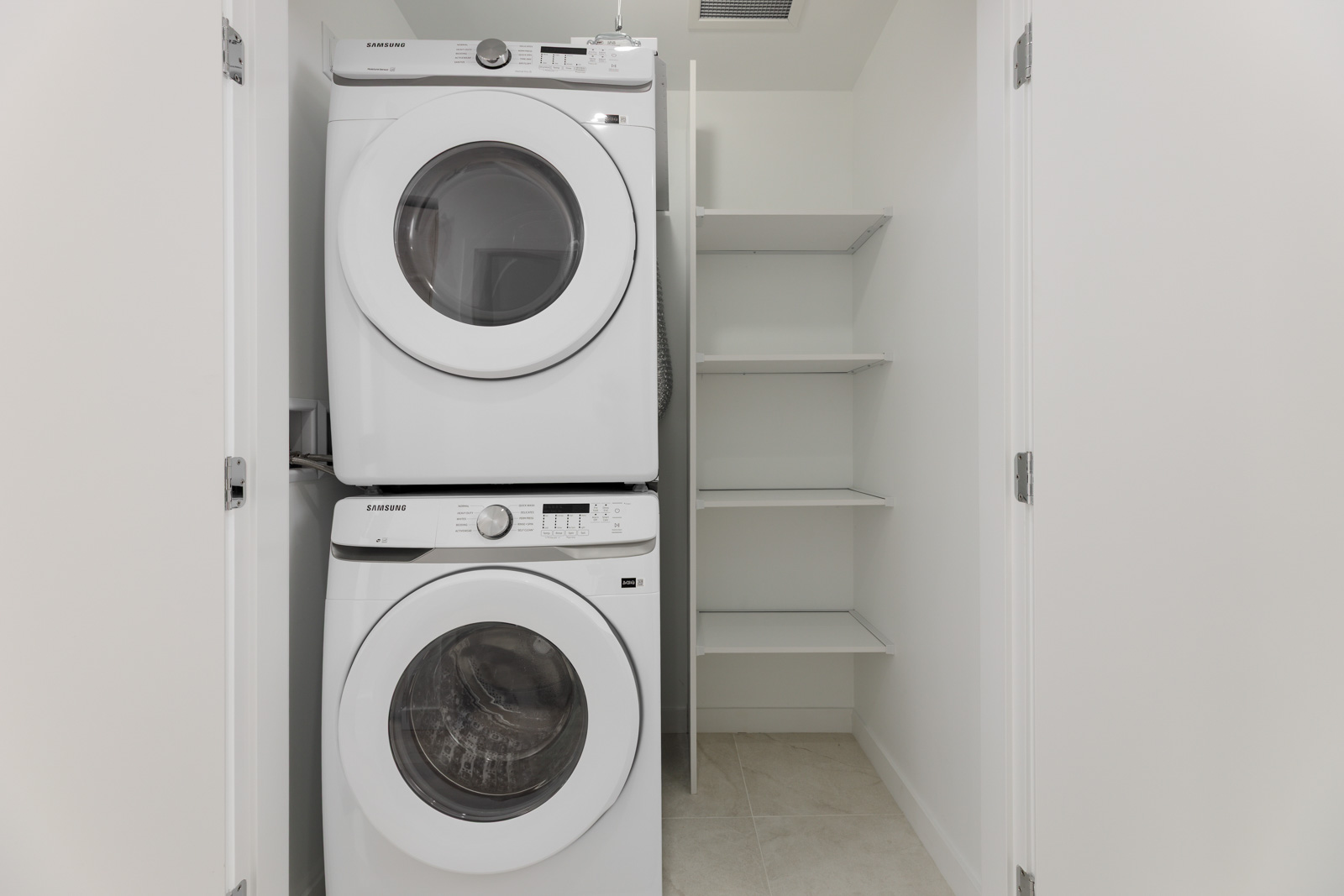 Stacked washer and dryer in a white laundry room with empty shelves on the side.