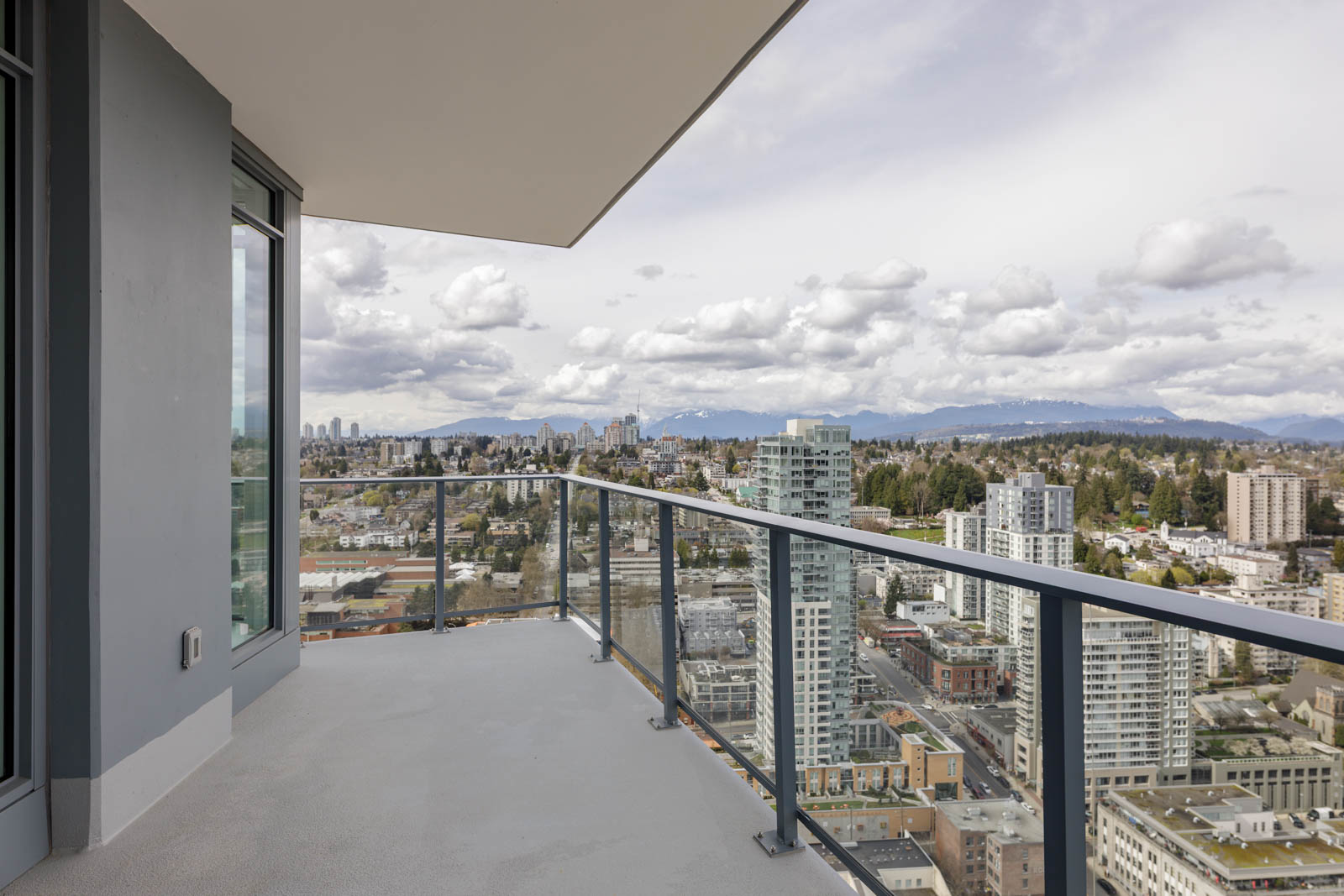 High-rise balcony with glass railing overlooks an urban landscape, featuring numerous buildings and a distant mountain range under a cloudy sky.
