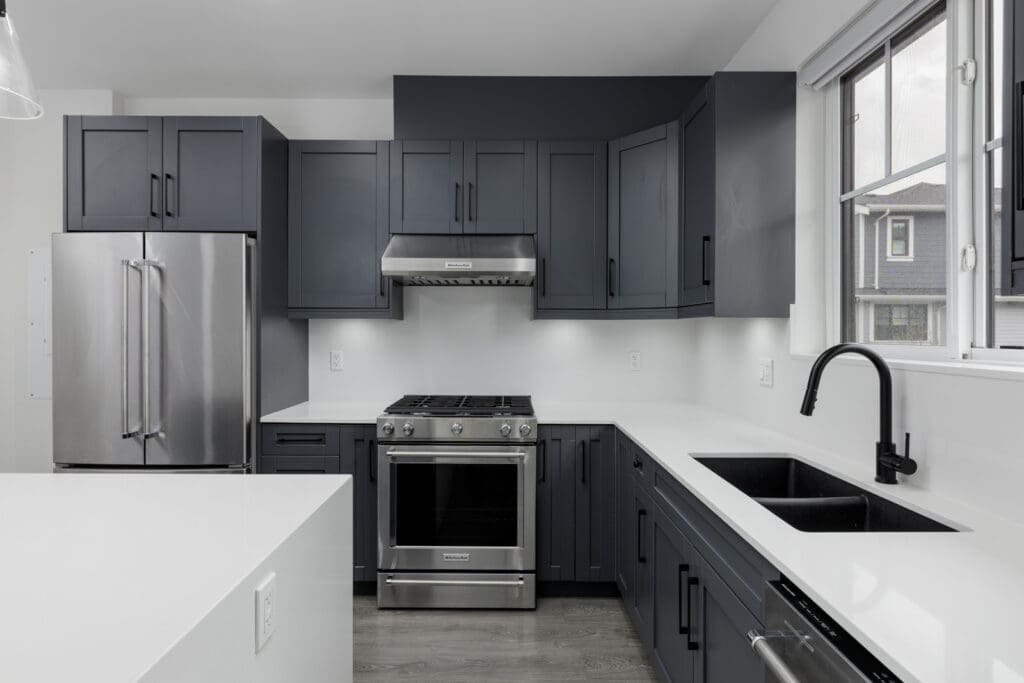 Modern kitchen with dark gray cabinets, stainless steel appliances, white countertops, and a double sink beneath a window.