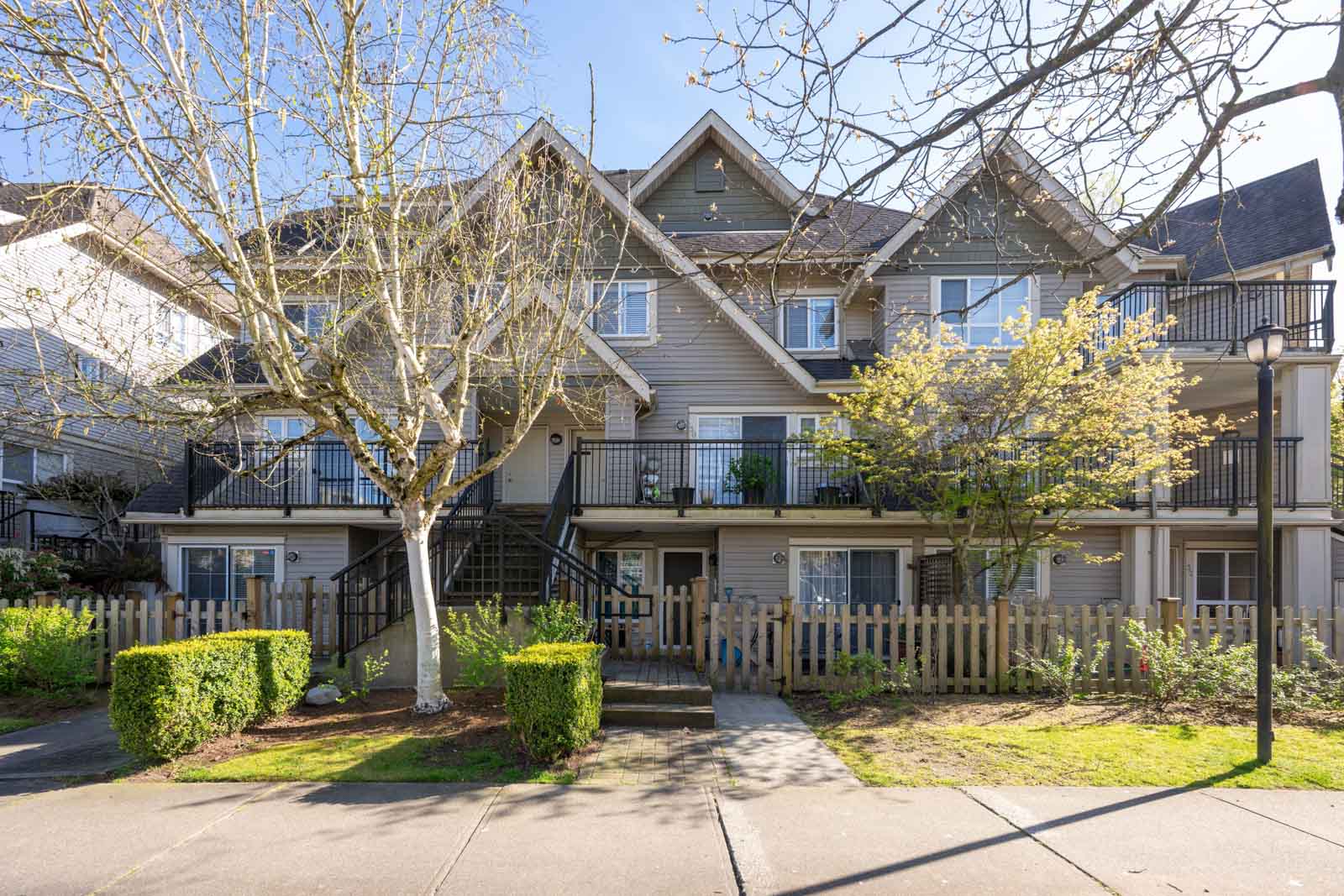 A multi-story townhouse building with balconies, a wooden fence, front steps, and trees in the yard on a sunny day.