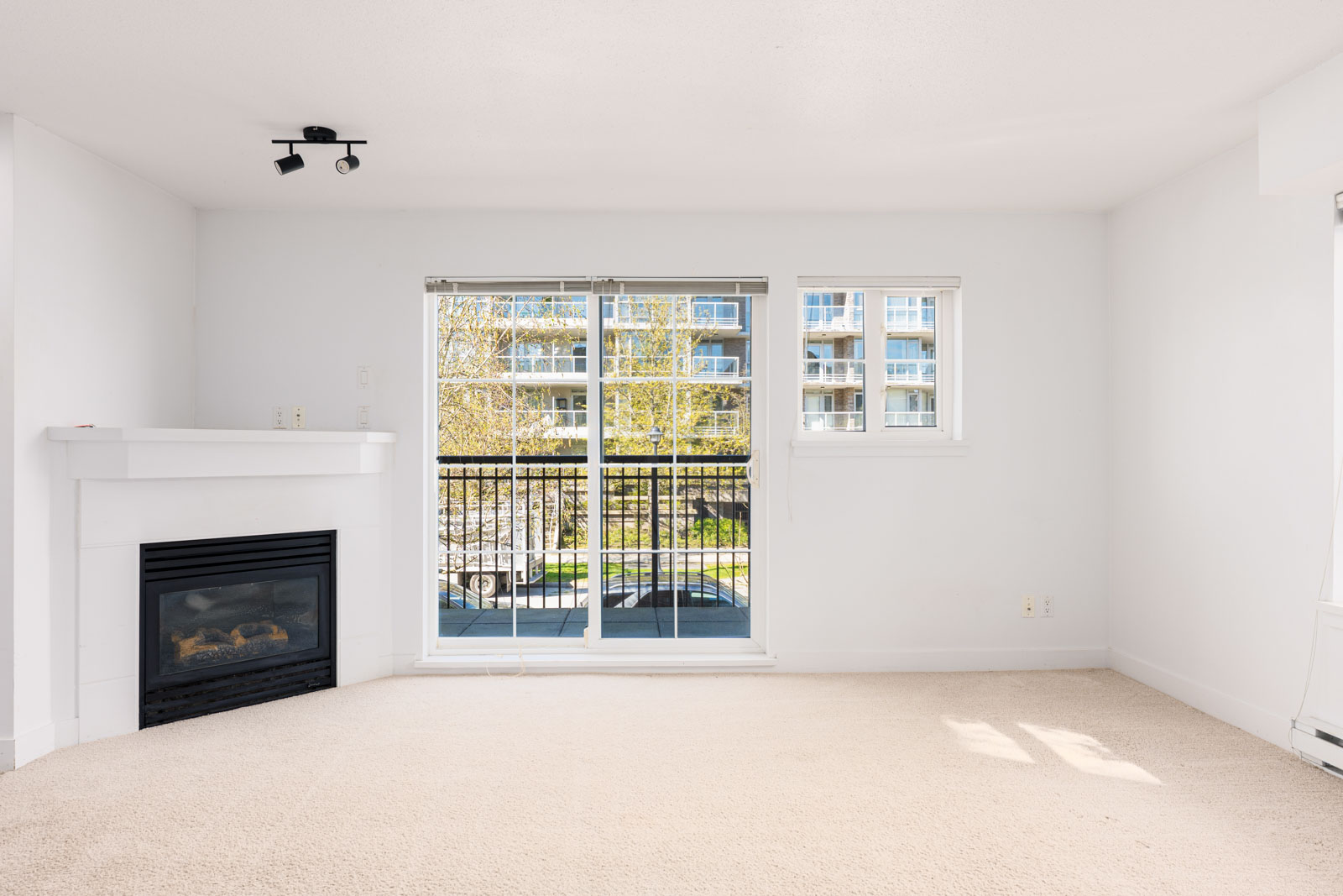 Unfurnished living room with a beige carpet, corner fireplace, and sliding glass doors leading to a balcony with a view of an apartment building.