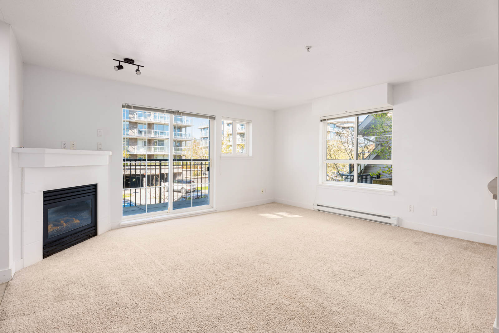 Unfurnished living room with beige carpet, white walls, fireplace, large windows, and a sliding glass door leading to a balcony with view of nearby apartment buildings.