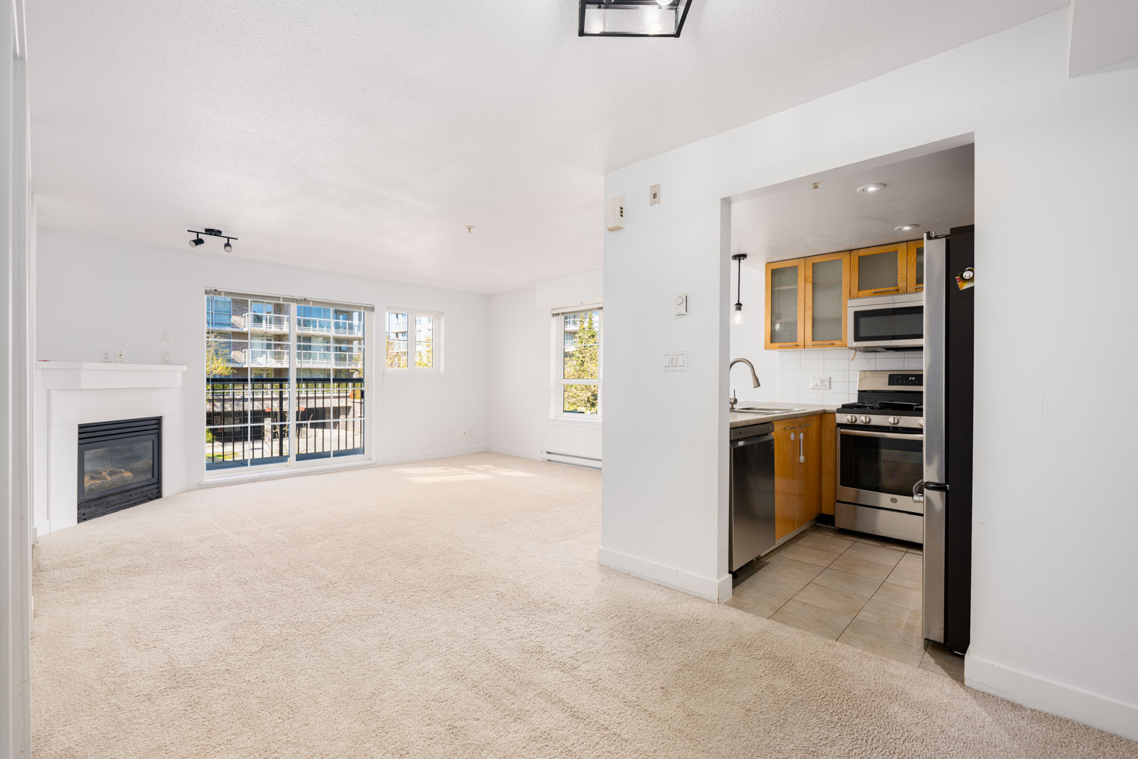 Bright, unfurnished apartment with an open living area, fireplace, carpeted floors, balcony access, and a small kitchen with stainless steel appliances visible to the right.