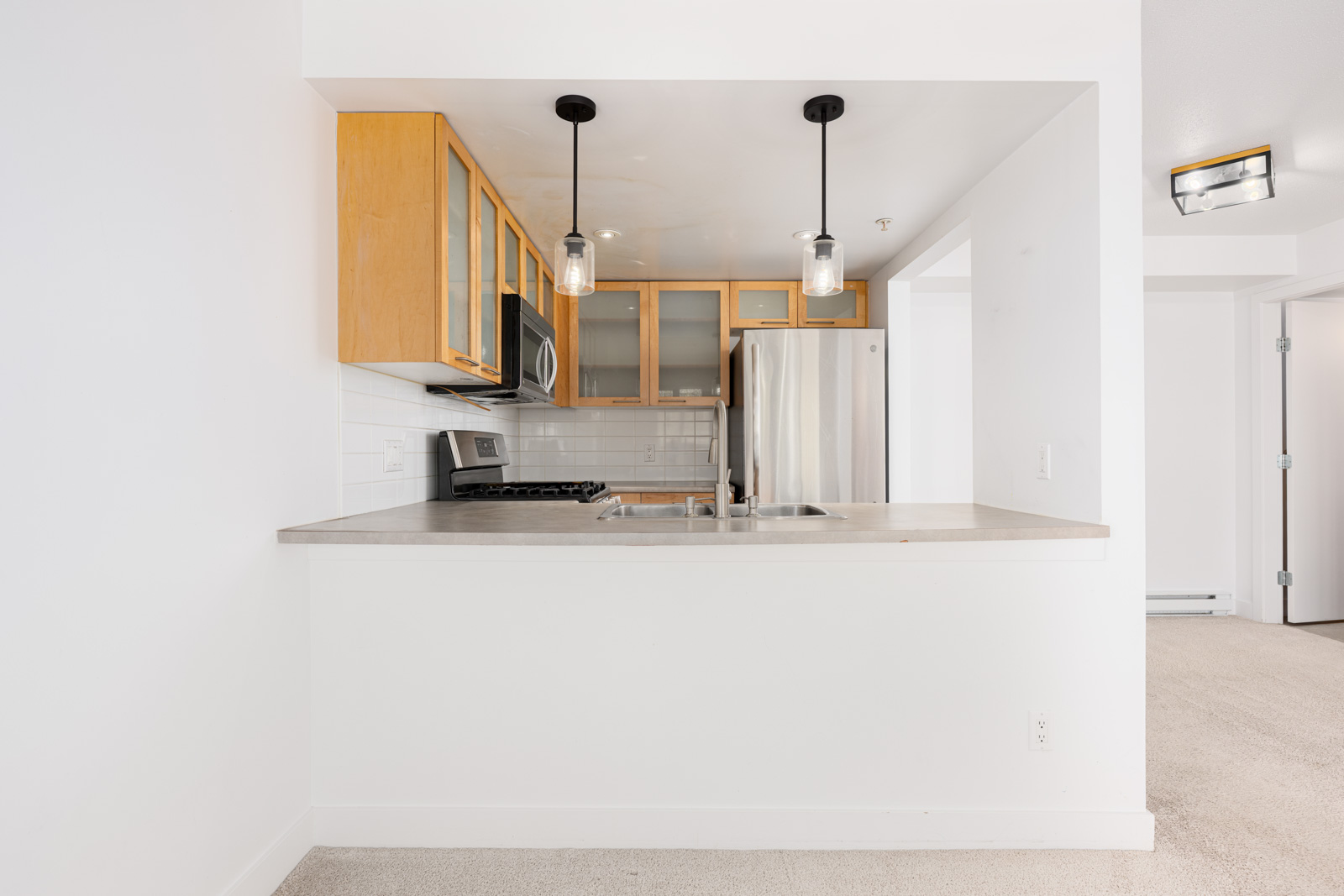 Modern kitchen with light wood cabinets, stainless steel appliances, two pendant lights, and a neutral countertop with an integrated sink.