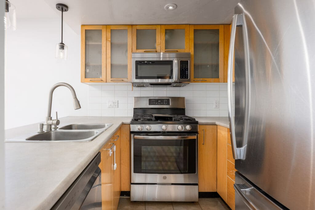 Compact modern kitchen with stainless steel appliances, light wood cabinets, a double sink, and a gas stove beneath a microwave.