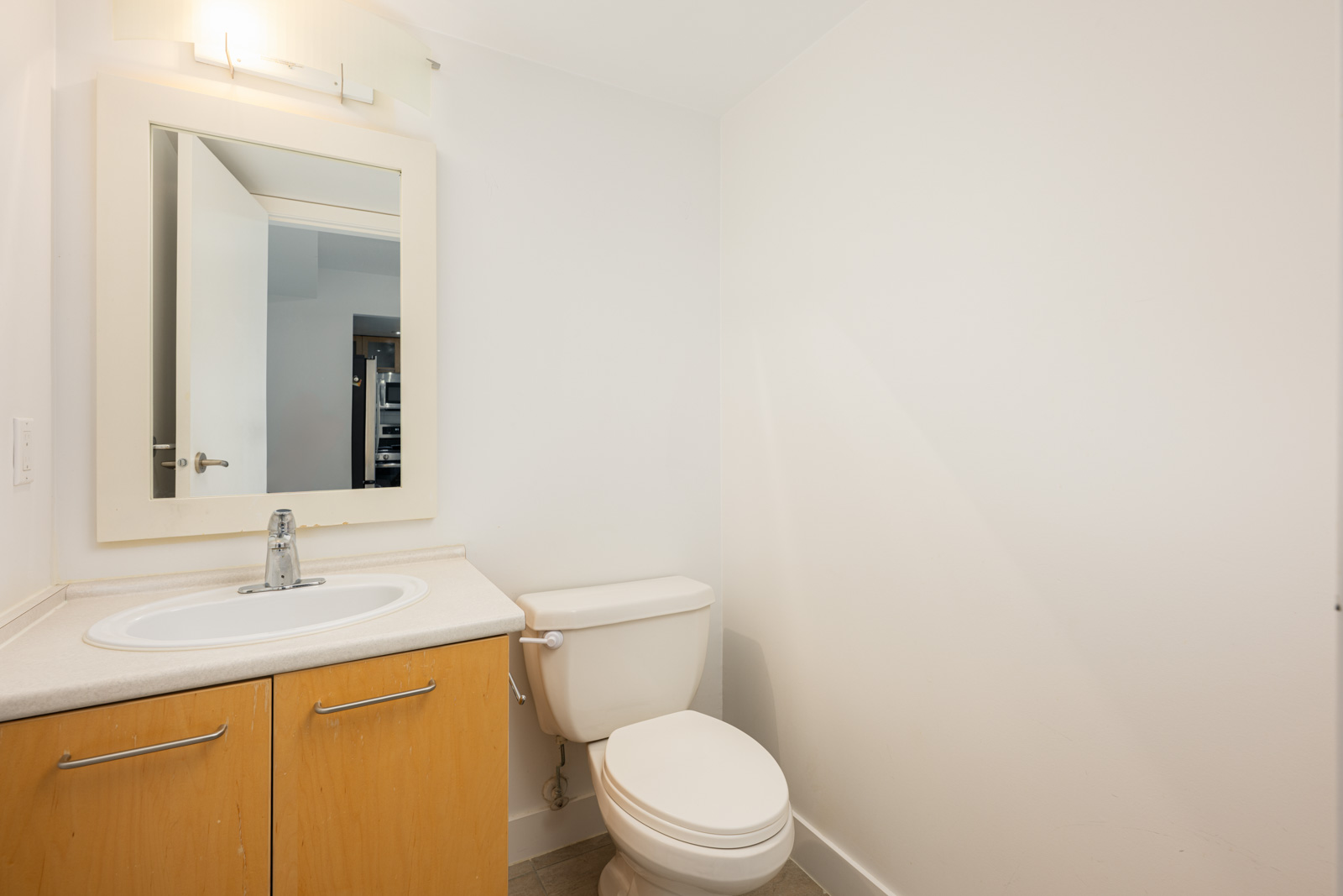 A small bathroom with a wall-mounted mirror, a single sink with a faucet, wooden cabinet, and a white toilet, all against plain white walls.