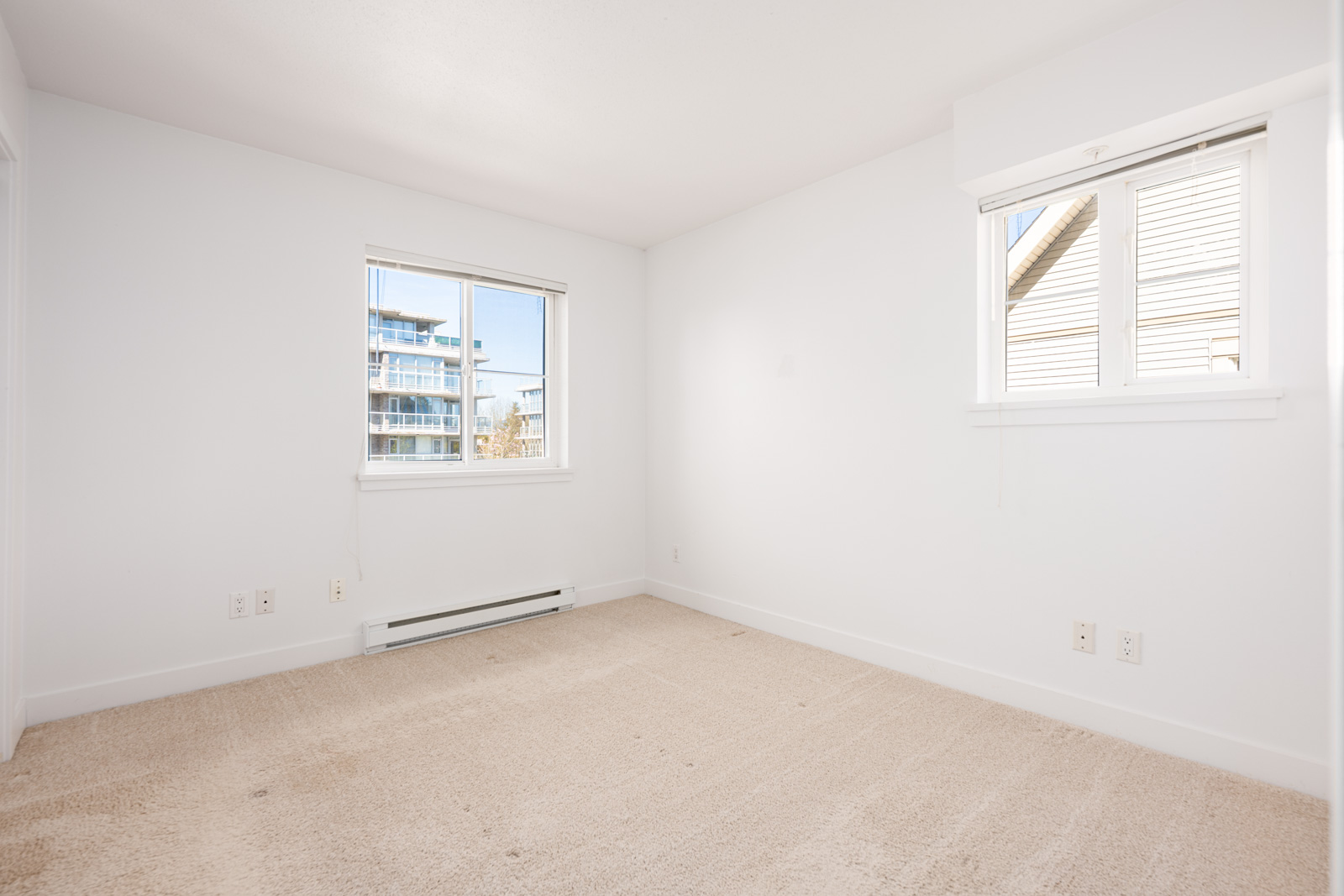 Empty room with beige carpet, white walls, two windows, baseboard heater, and multiple wall outlets; sunlight coming in through the windows.