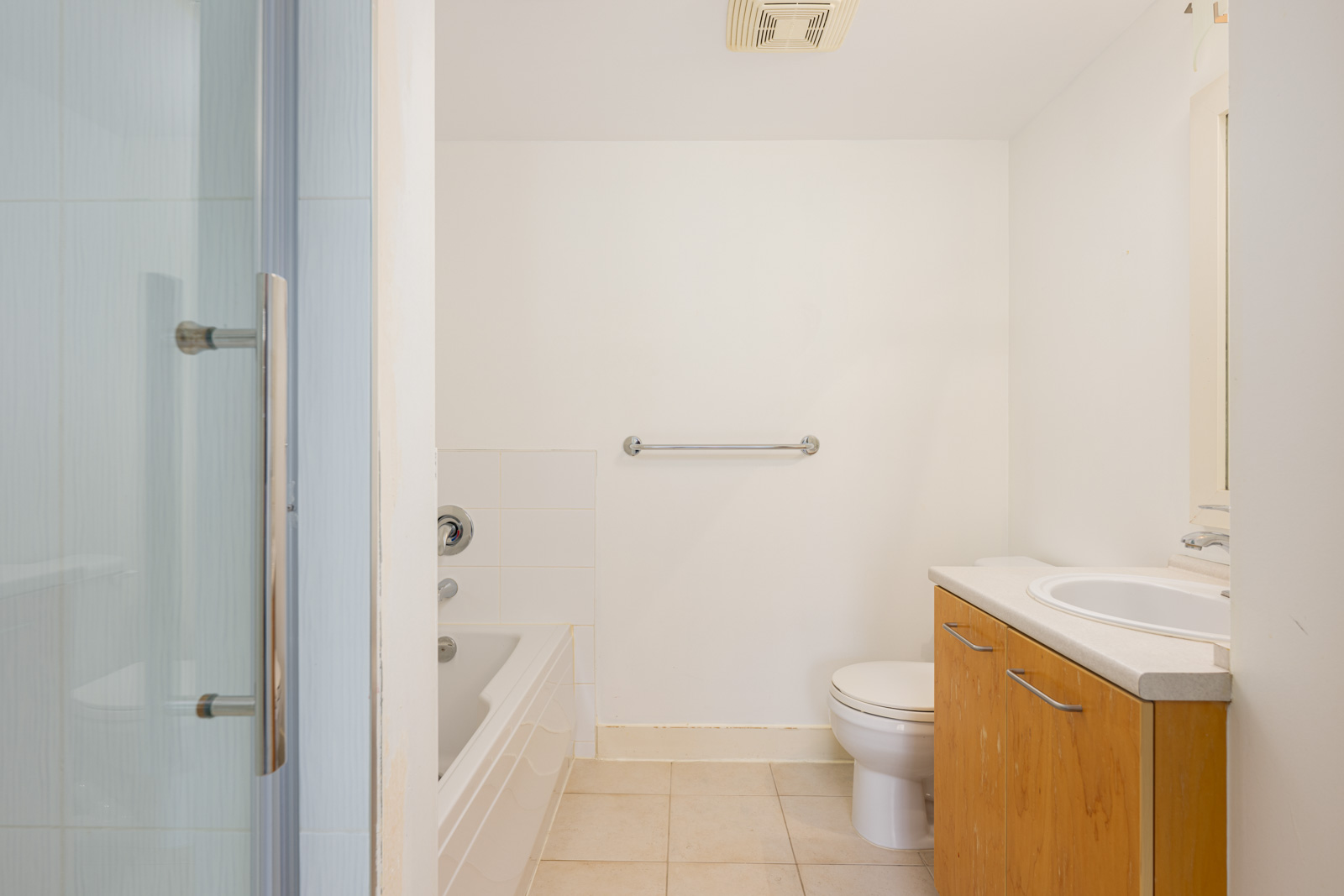 A small bathroom with a bathtub, toilet, sink with a wooden cabinet, grab bar, and light-colored tiled floor.
