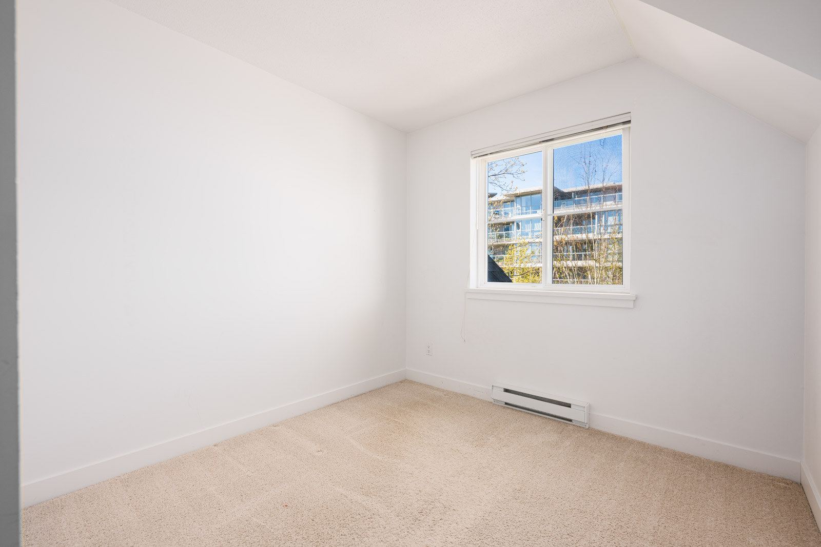 Empty room with white walls, beige carpet, a window with blinds partially open, and a baseboard heater below the window.