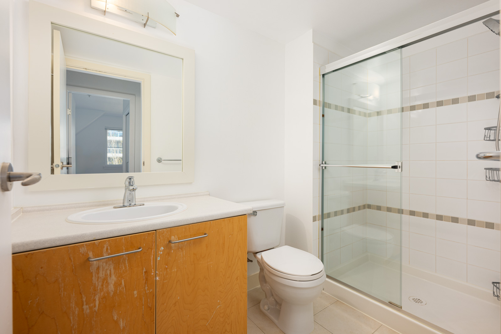 Bathroom with a sink, wooden cabinet, toilet, and a glass-enclosed shower. The walls and tiles are white, and a mirror is above the sink.