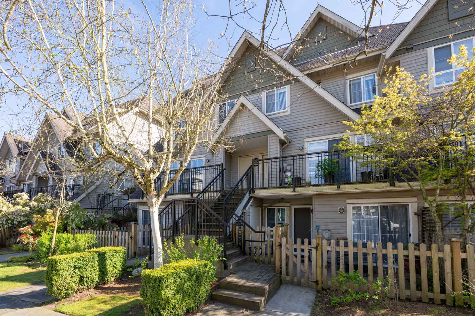 A row of modern townhouses with front porches, balconies, picket fences, and landscaped gardens on a sunny day.