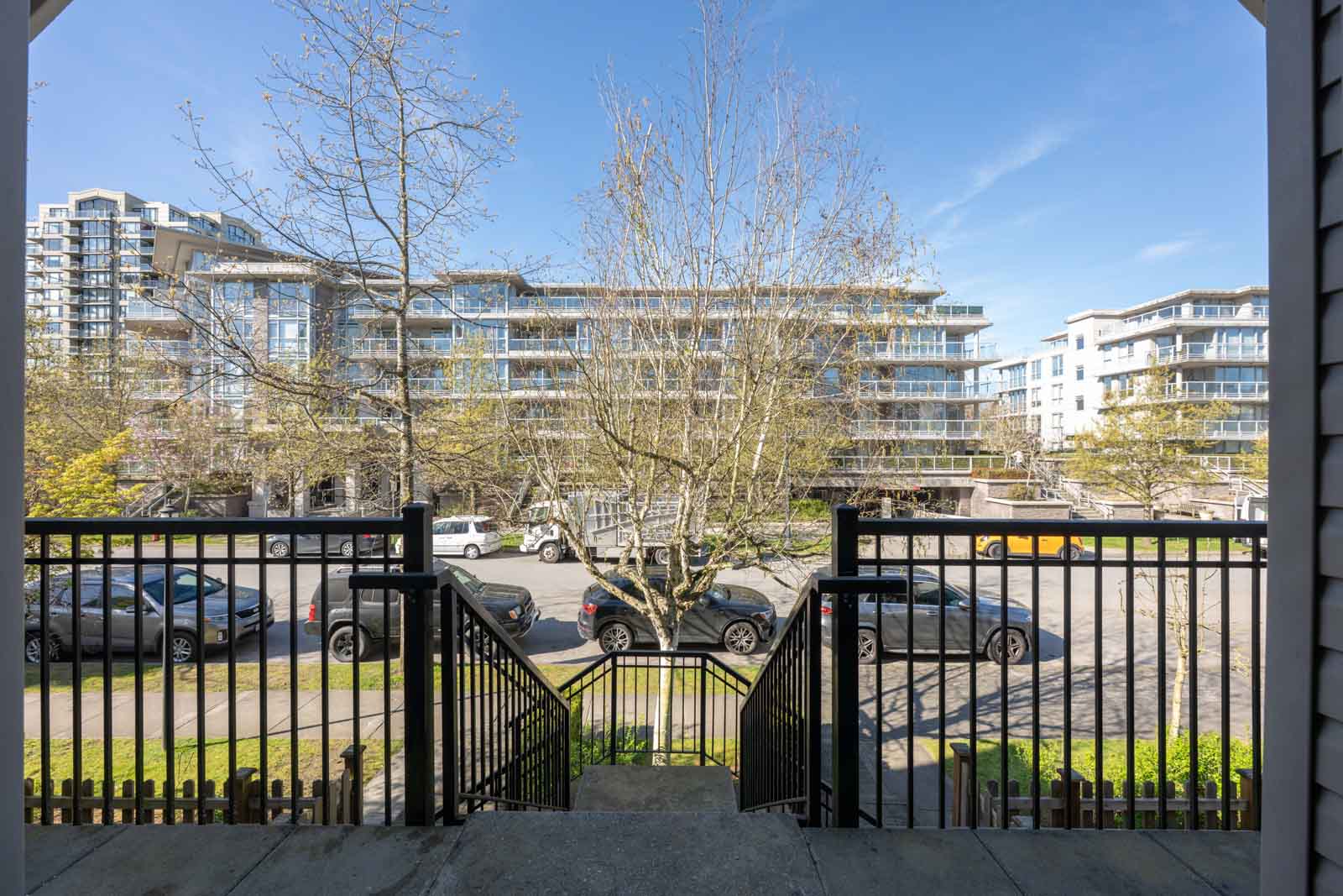 View from a porch showing steps leading down to a sidewalk, parked cars, bare trees, and modern apartment buildings in the background on a clear day.
