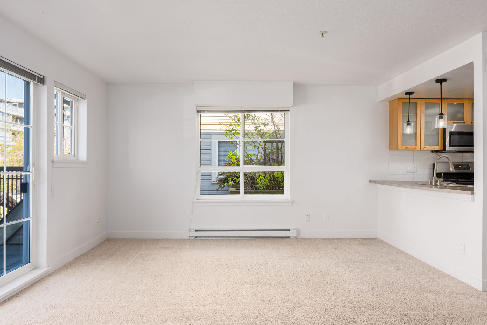 Unfurnished living room with beige carpet, large window, glass door to balcony, and open view into kitchen with wood cabinets and pendant lights.