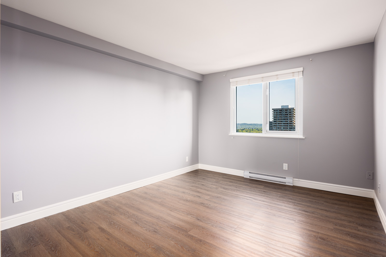 Empty room with light grey walls, a large window with a view of buildings and trees, and wood laminate flooring.