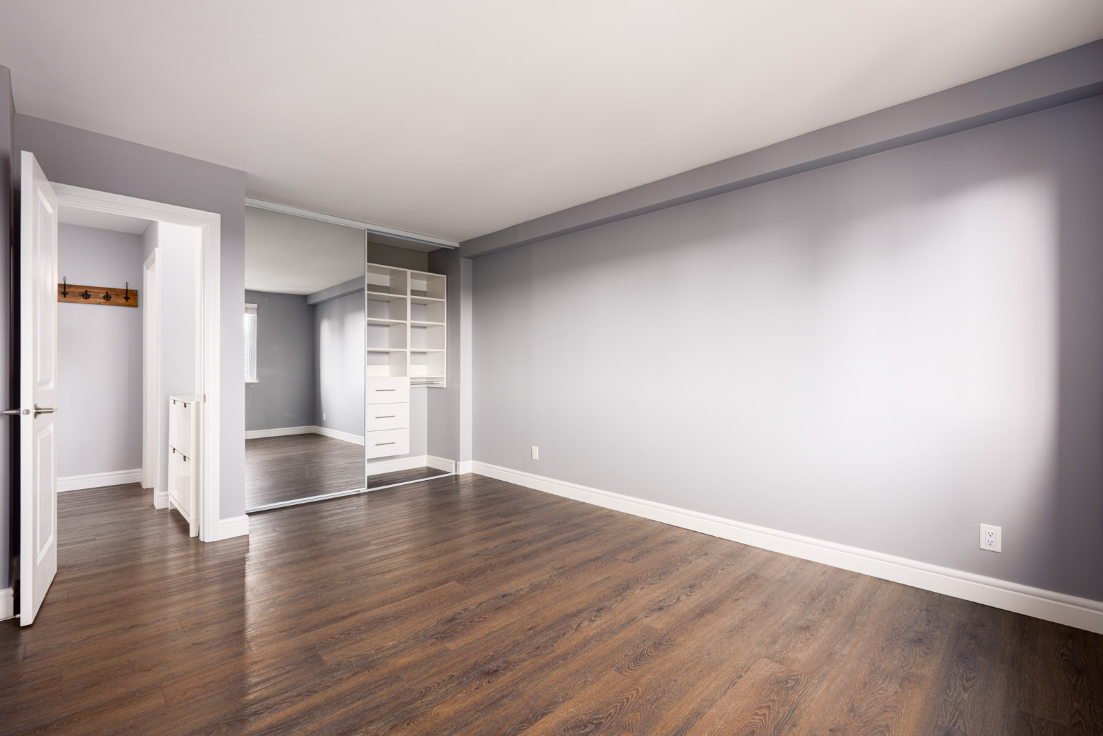 Empty room with gray walls, dark wood flooring, a large mirrored closet, open white door, and built-in shelving inside the closet.