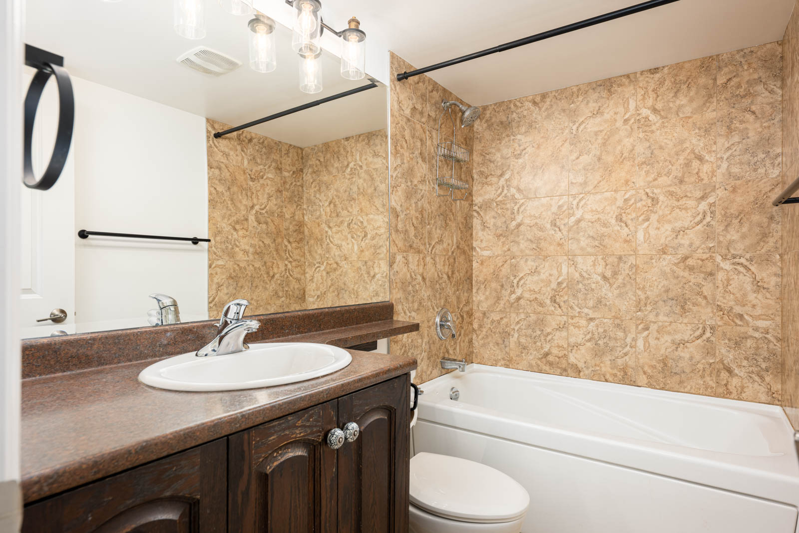 Bathroom with a brown countertop, white sink, dark wood vanity, white bathtub, and tan tiled shower area. Towel rack and mirror are mounted on the wall.
