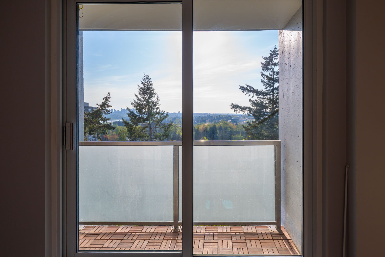 A view through a glass sliding door reveals a small balcony with wood-patterned flooring, frosted railing, and trees in the distance under a clear sky.