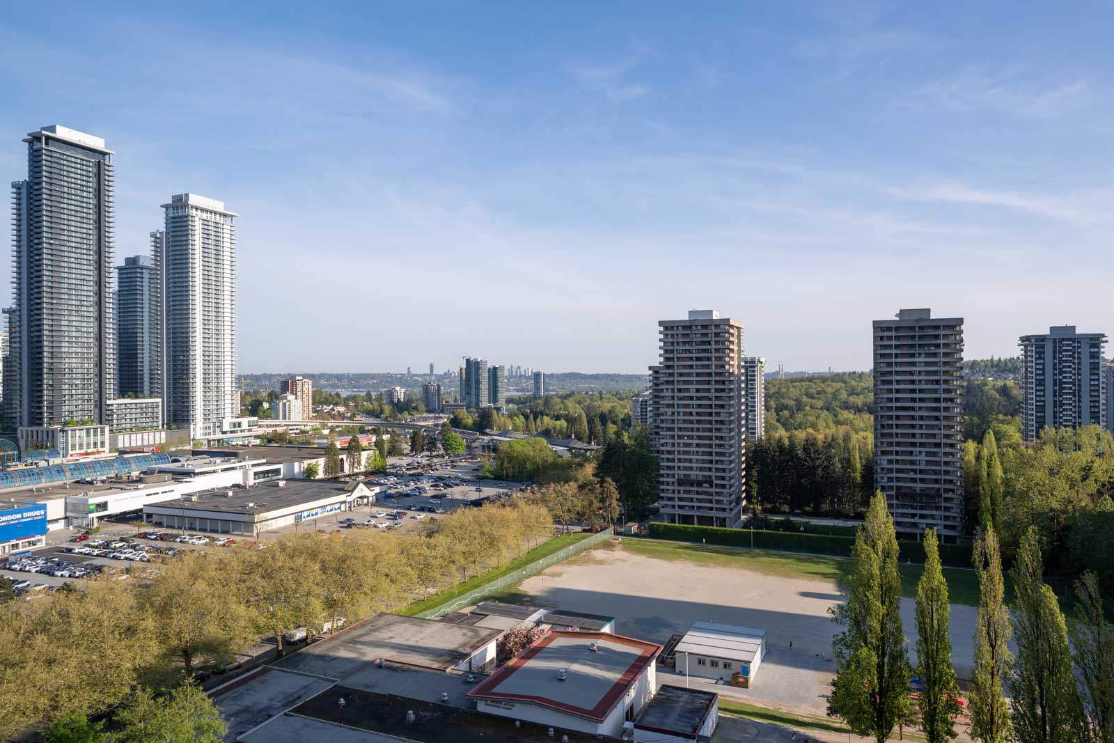 A cityscape showing high-rise apartment buildings, a large parking lot, a sports field, and trees under a clear blue sky.