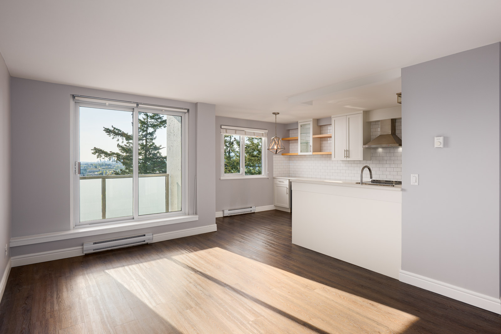 Bright, modern apartment with large windows, open kitchen, white cabinets, stainless steel appliances, and wood flooring. Sliding door leads to a balcony with a scenic view.