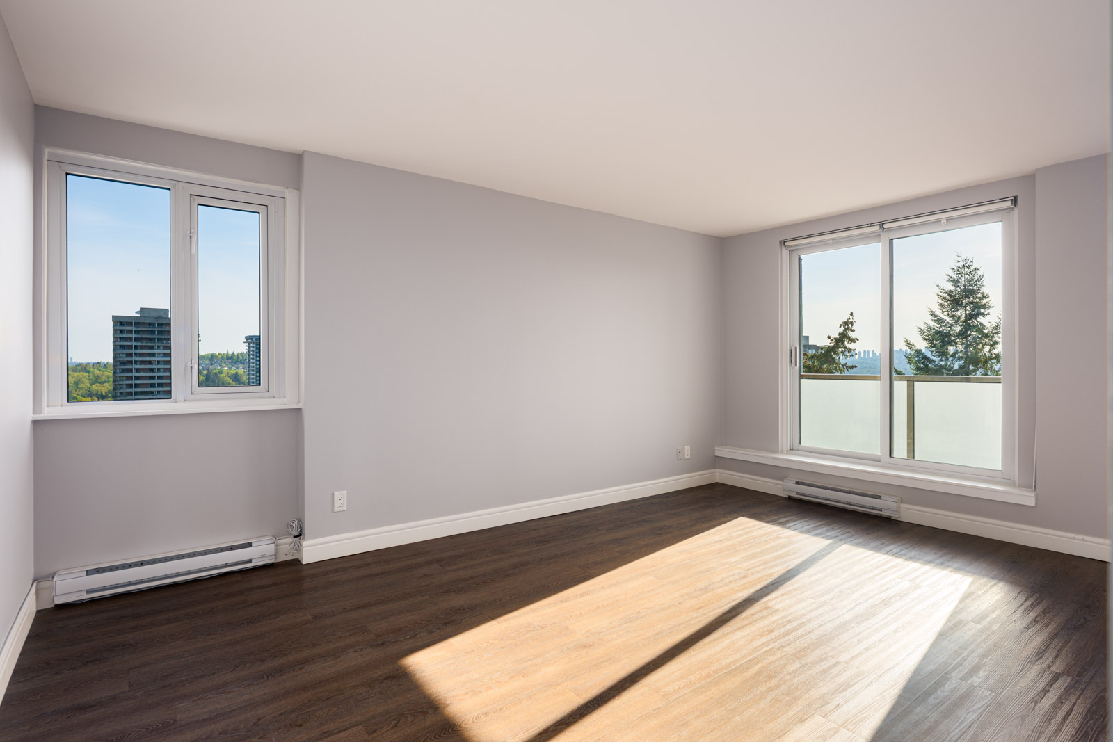 Empty room with light gray walls, large windows, sliding glass door to balcony, and dark wood flooring. Natural light fills the space, with views of trees and buildings outside.