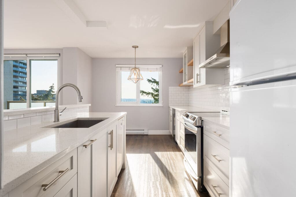 Bright modern kitchen with white cabinets, stainless steel appliances, a central island with a sink, and large windows letting in natural light.